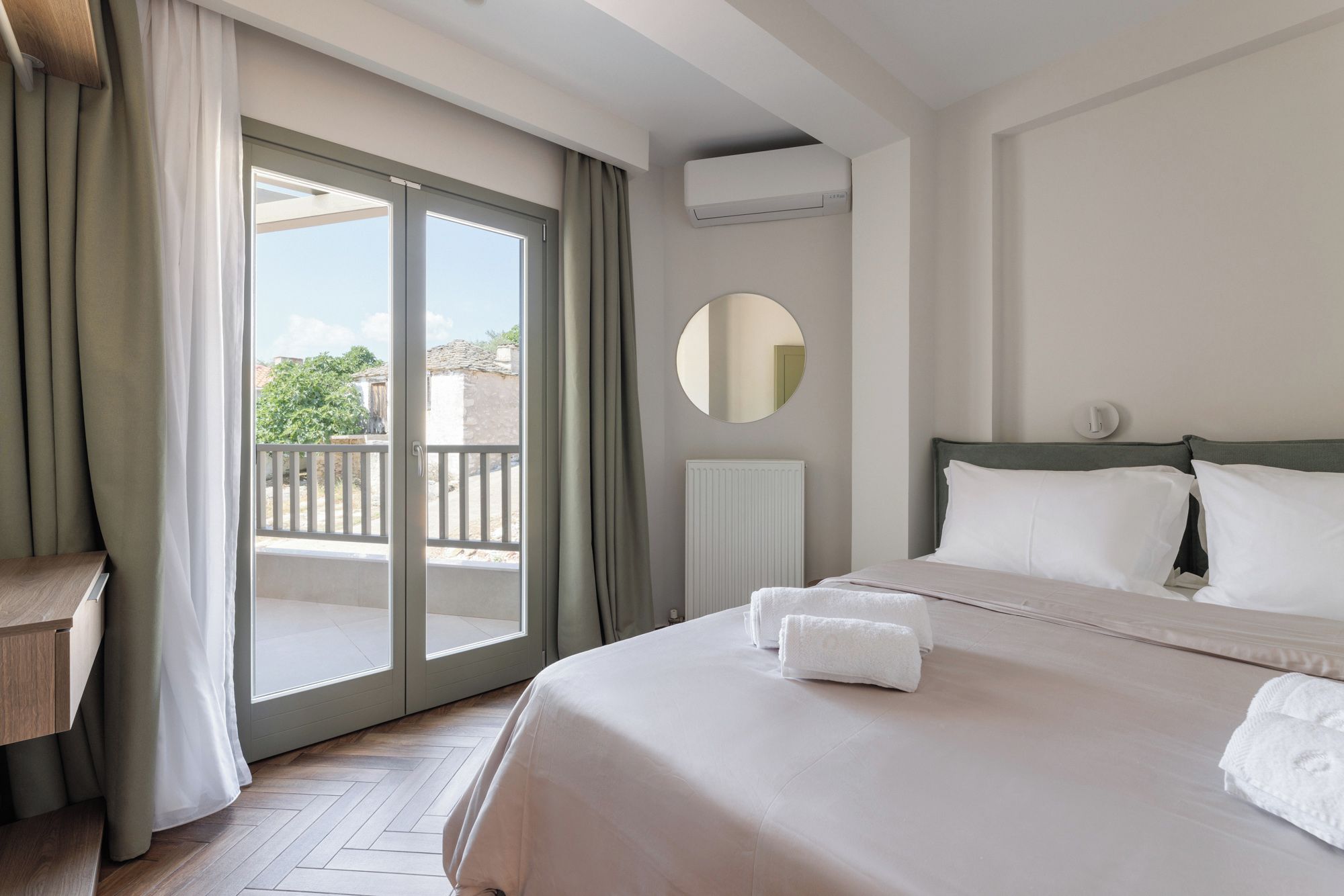 A hotel room with a bed, a round mirror, a radiator, and a view of the outdoors.