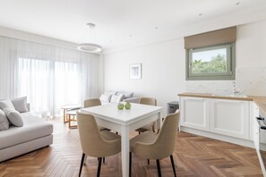 A modern living room with a sofa, dining table, and chairs. There is a kitchen area with cabinets and a window.