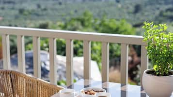 A table with two cups of coffee and a bowl of cookies, a potted plant, wicker chairs, and a view of a green landscape.