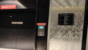 Um elevador com mostrador digital, uma placa vermelha acima da porta e um painel fixado na parede com texto.