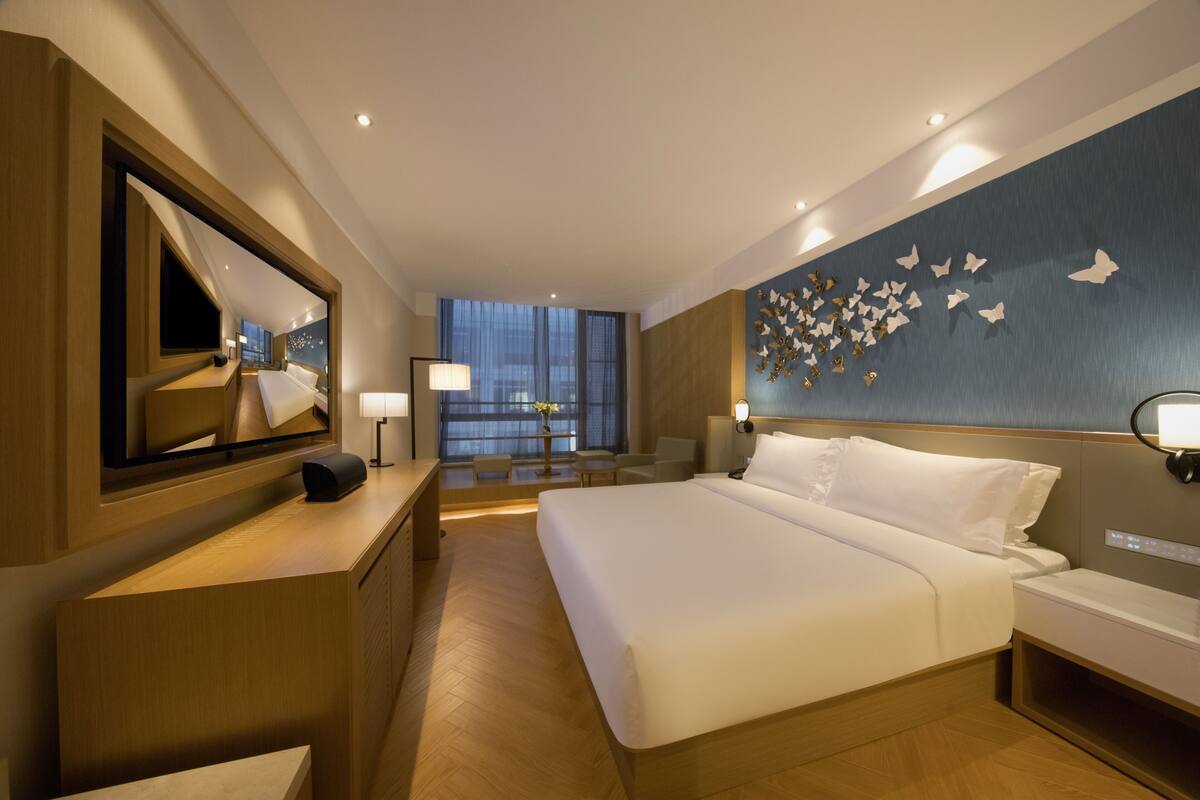 A hotel room with a large bed, a desk with a computer, a mirror, a wall with butterfly decorations, and a view of the city at night.