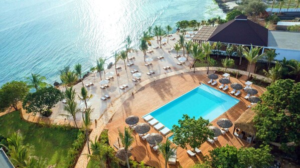 An aerial view of a resort with a swimming pool surrounded by sun loungers and umbrellas, a beach area with lounge chairs, and palm trees.