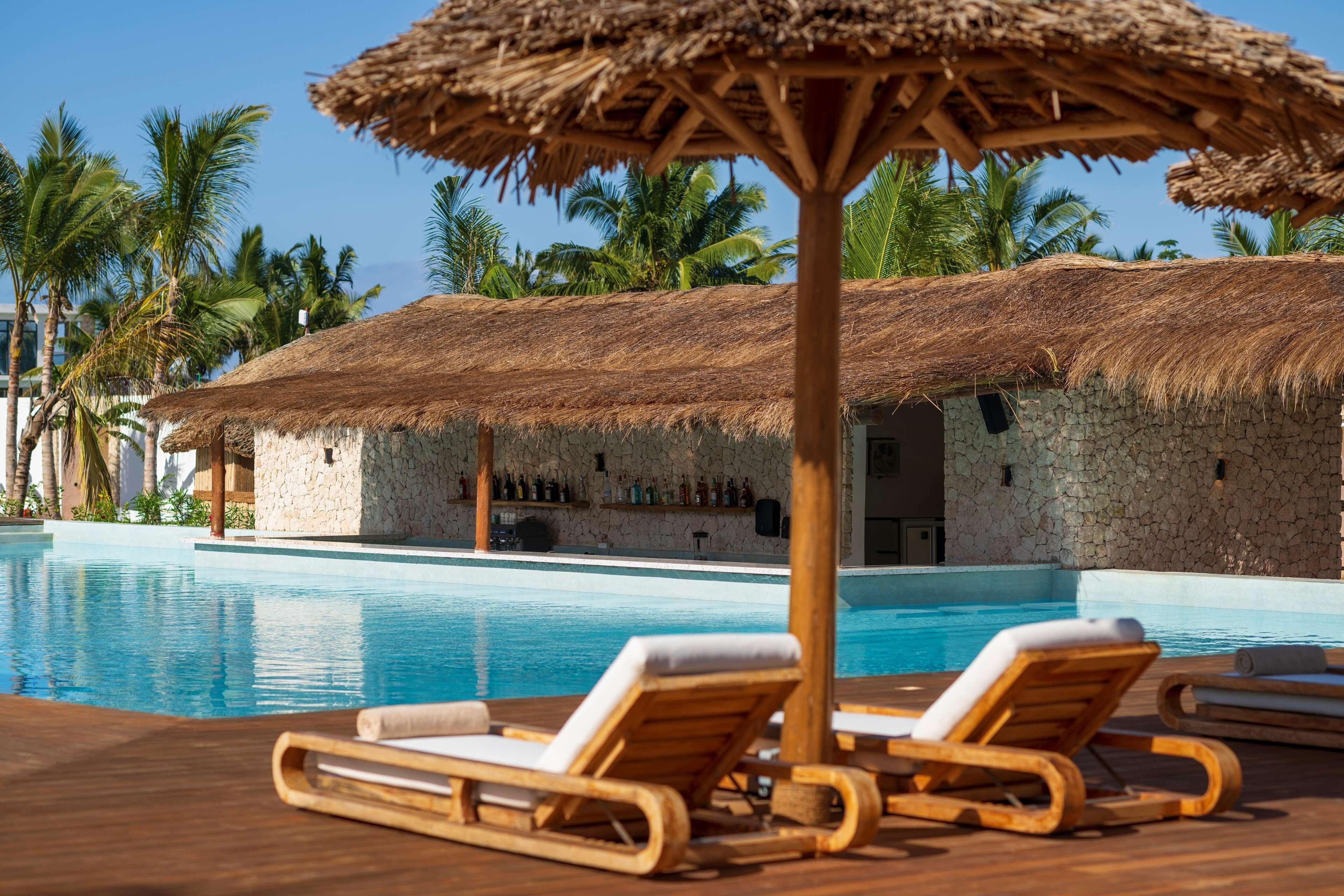 Un espace aménagé au bord de la piscine, avec des chaises longues, une structure à toit de chaume et un bâtiment en pierre en arrière-plan.