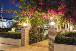 Un espacio exterior bien iluminado con un pilar de ladrillo, una pérgola metálica con flores rosas y un sendero pavimentado.
