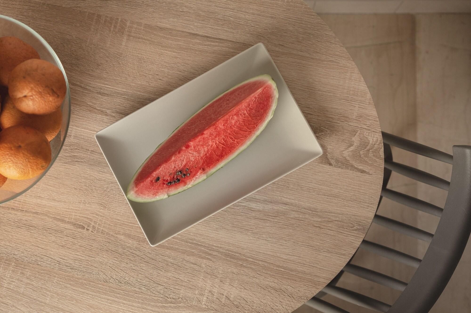 A round wooden table with a white plate containing a slice of watermelon and a bowl of oranges.