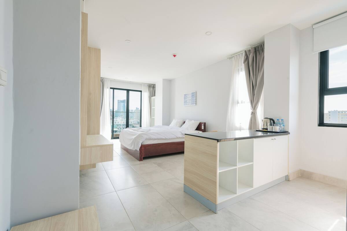 A modern hotel room with a bed, a kitchenette, a large window, and a city view.