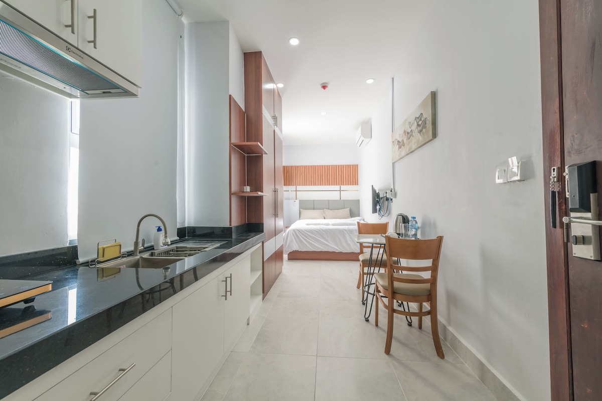 Una cocina moderna con encimera negra, armarios blancos y estufa integrada. Hay una zona de comedor con una mesa y sillas, y una cama con cabecero de madera al fondo.
