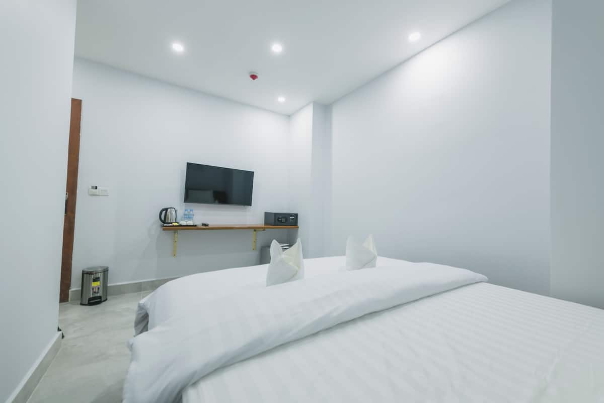 A modern hotel room with a large bed, a flat-screen TV mounted on the wall, a small shelf with a kettle and water bottles, and a trash bin.