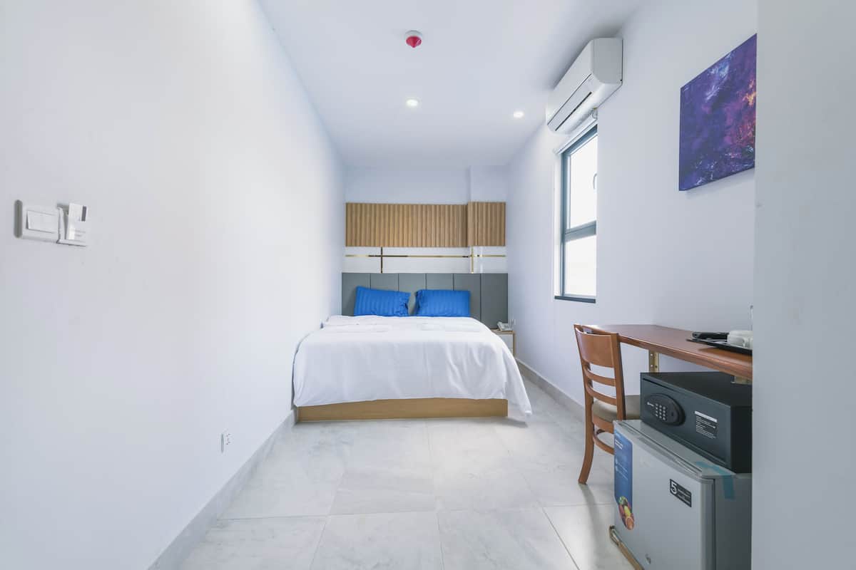 A bedroom with a bed, a desk, a chair, a small refrigerator, and a window.