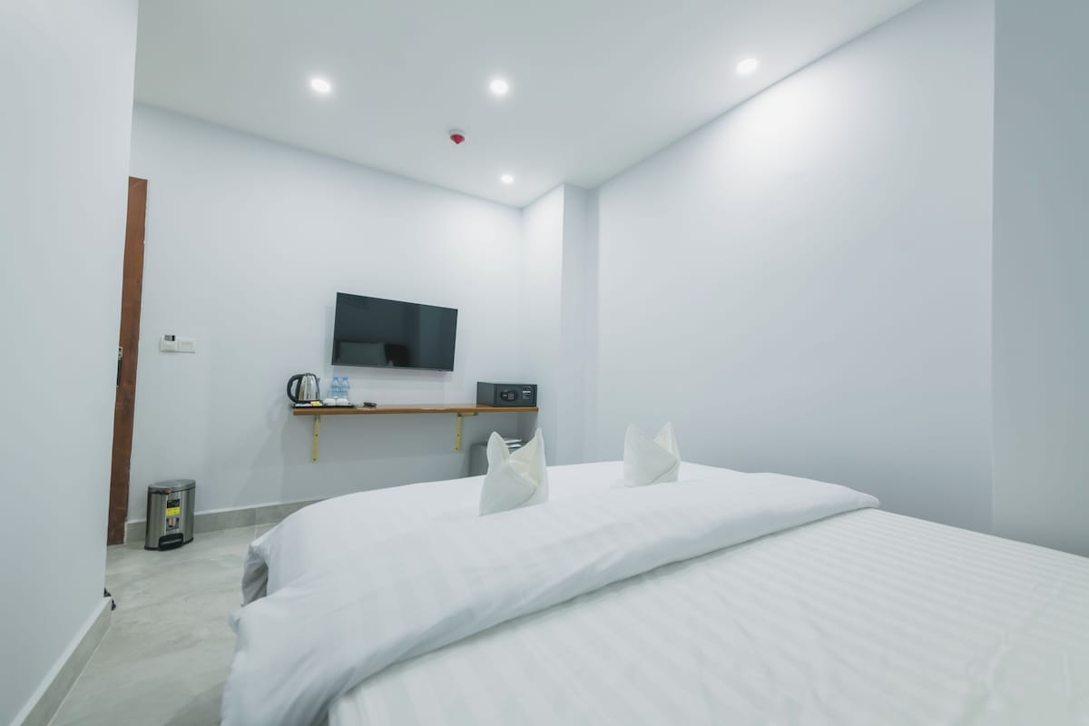 Una habitación de hotel moderna con una cama grande, un televisor de pantalla plana en la pared, un estante pequeño con una tetera y botellas de agua, y un cesto de basura.