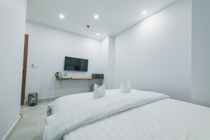 Ein modernes Hotelzimmer mit einem großen Bett, einem flachbildfernseher an der Wand, einem kleinen Schrank mit Wasserkocher und Wasserflaschen sowie einem Mülleimer.