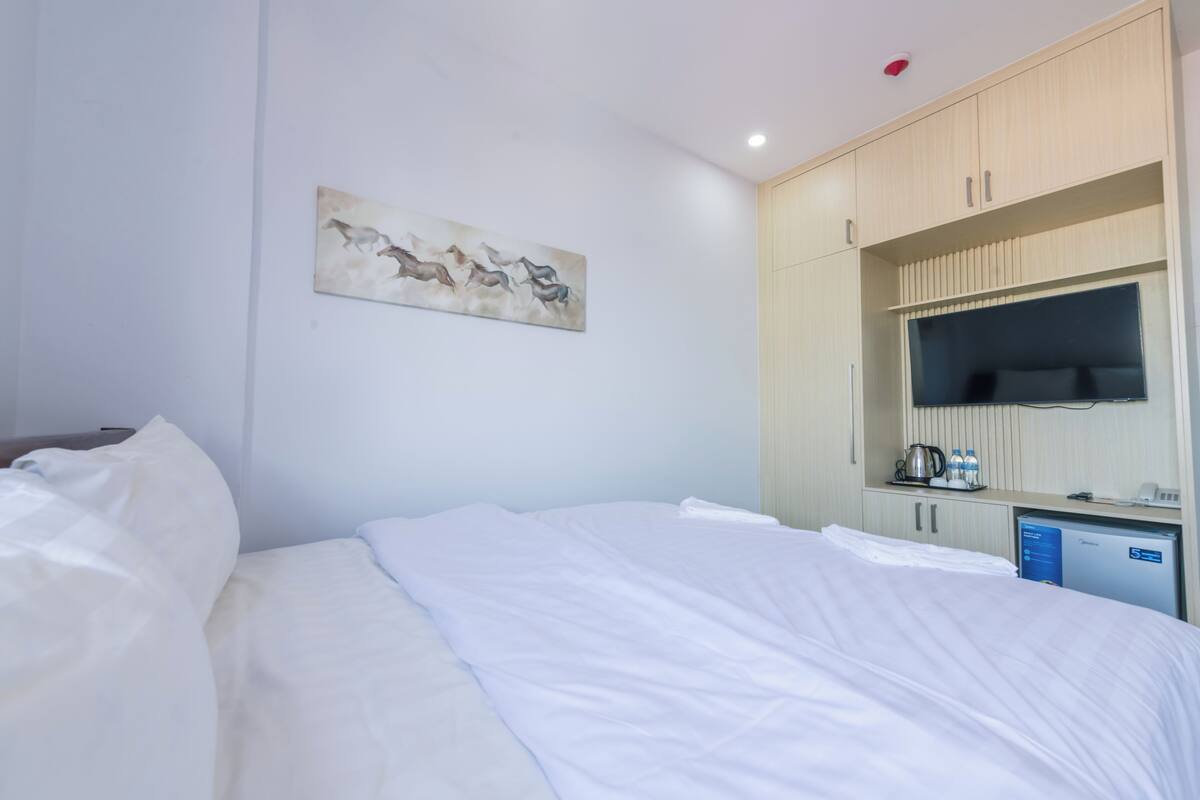 A hotel room with a bed, a painting on the wall, a TV, and a minibar.