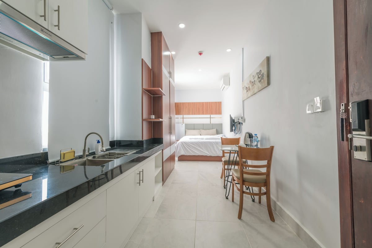 A modern kitchen with a black countertop, white cabinets, and a built-in stove. There is a dining area with a table and chairs, and a bed with a wooden headboard in the background.