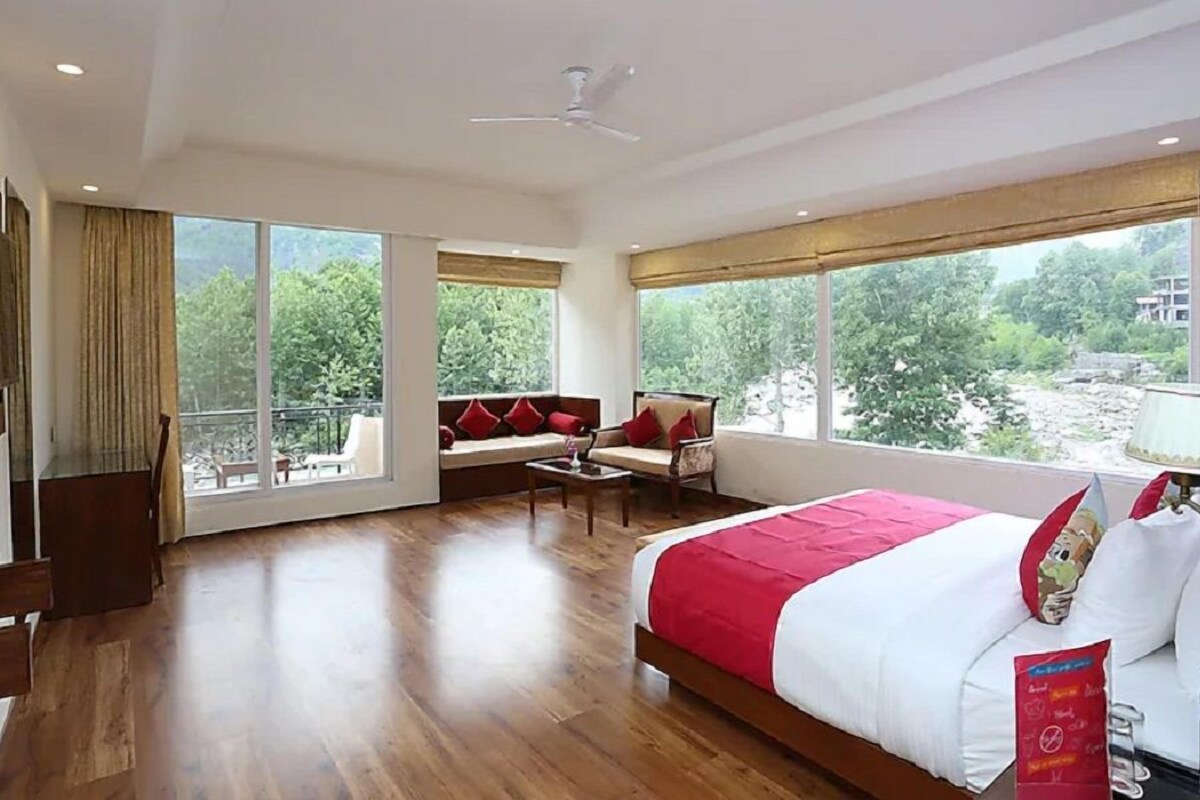 A hotel room with a large bed, a sofa, a small table, and a balcony with a view of greenery.
