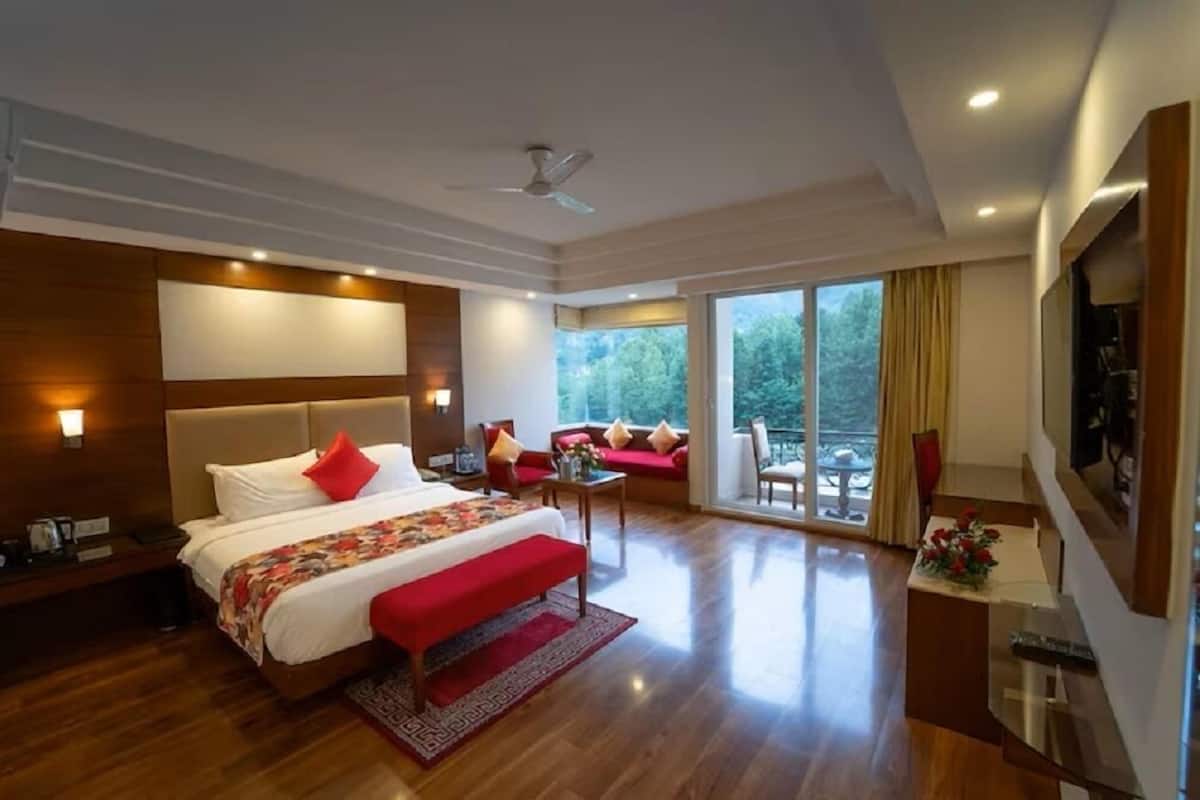 A modern hotel room with a large bed, a sofa, a TV, and a balcony with a view of trees.
