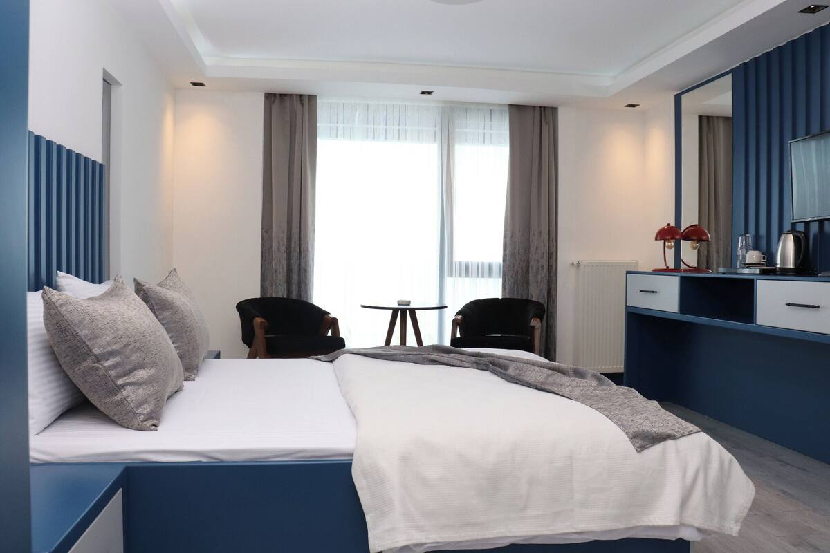 Moderni hotellihuone, jossa on sänky, kaksi tuolia, pieni pöytä, peili ja näkymä ulos.