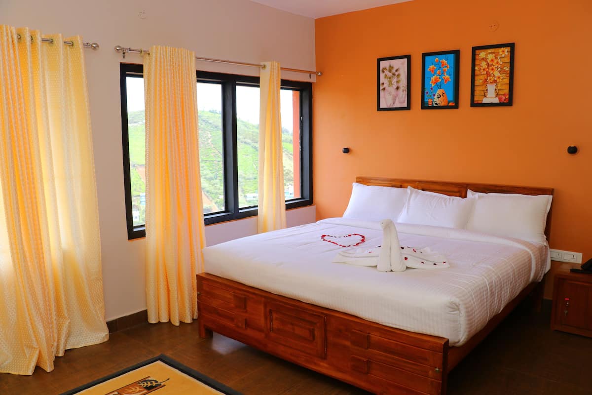 A neatly made bed with white linens and a heart-shaped decoration, a window with curtains, and three framed artworks on the wall.