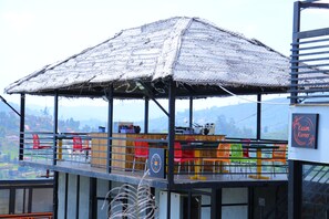 A restaurant with a thatched roof, outdoor seating with colorful chairs, and a view of the landscape. - The Frame Resorts Ooty (Coonoor)