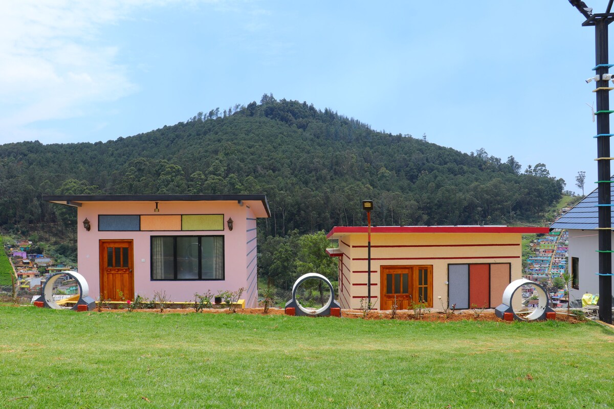 Two small buildings with large windows and doors, surrounded by grass and hills in the background.