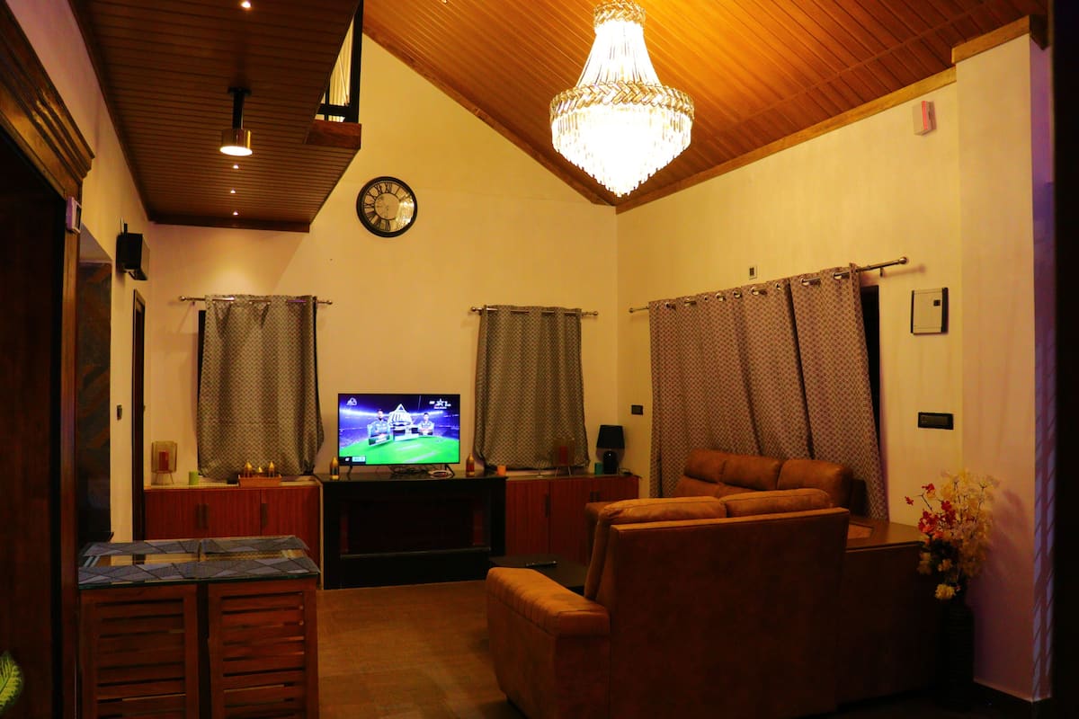A living room with a television, a chandelier, curtains, and a clock on the wall.