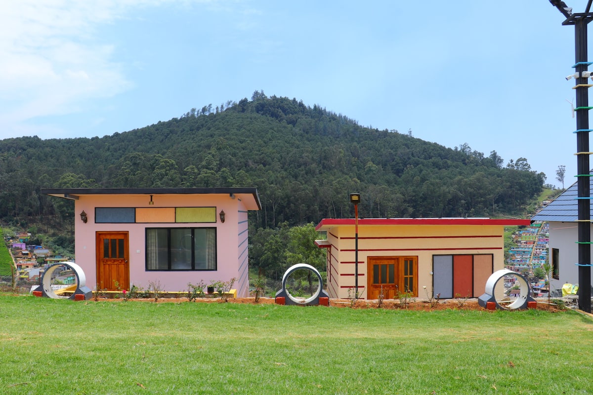 Two small buildings with large windows and doors, surrounded by grass and hills in the background.