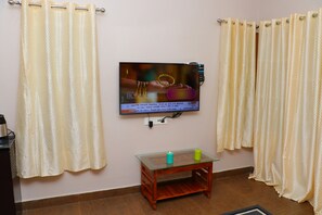 A living room with a flat-screen TV mounted on the wall, a glass coffee table with two candles, and curtains on the windows. - The Frame Resorts Ooty (Coonoor)