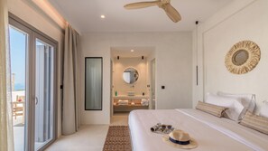 A modern hotel bedroom with a large bed, a ceiling fan, a round mirror, and a view of the beach through the sliding doors. - Nove Milos Luxury (Milos)