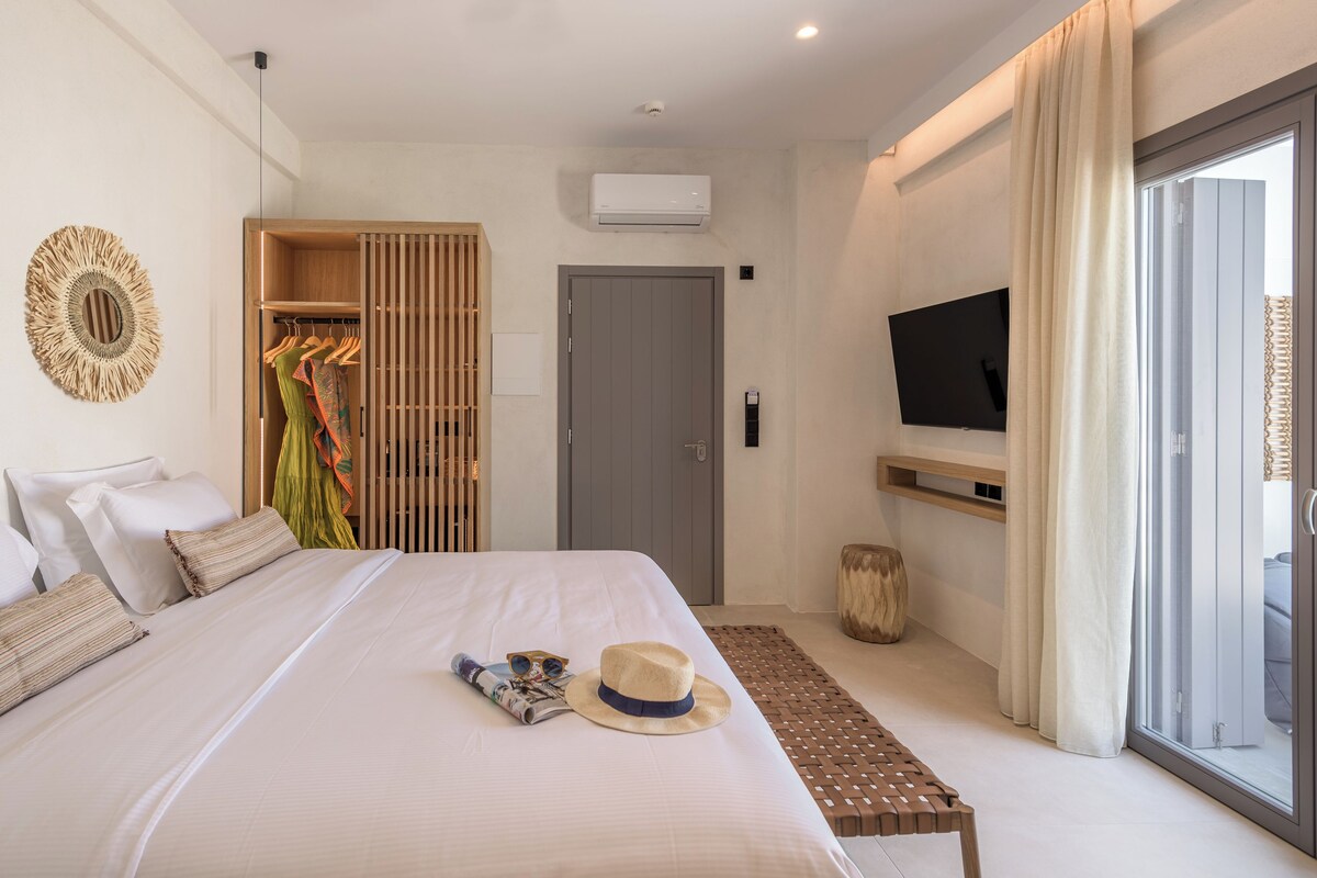 A modern hotel room with a large bed, a television mounted on the wall, and a sliding glass door leading to a balcony.