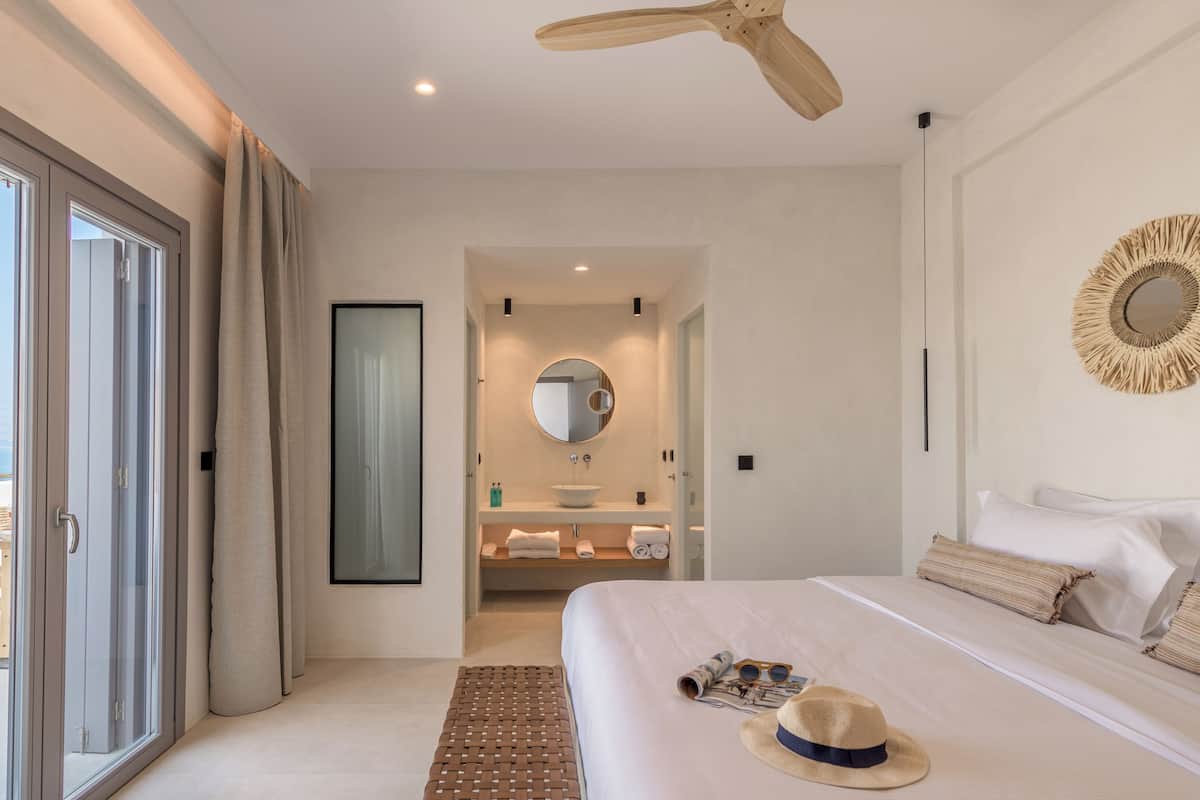 A modern hotel bedroom with a large bed, a ceiling fan, a round mirror, and a view of the beach through the sliding doors.