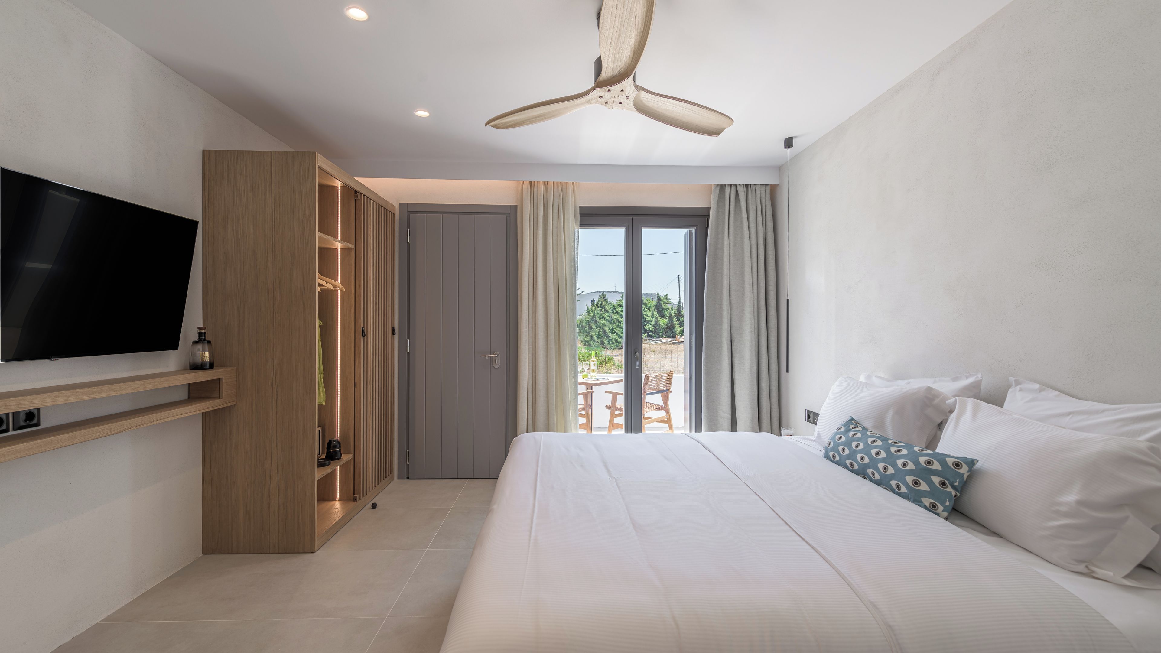 Quarto de hotel moderno com uma cama grande, televisão presa na parede, guarda-roupa e varanda com área de estar ao ar livre.