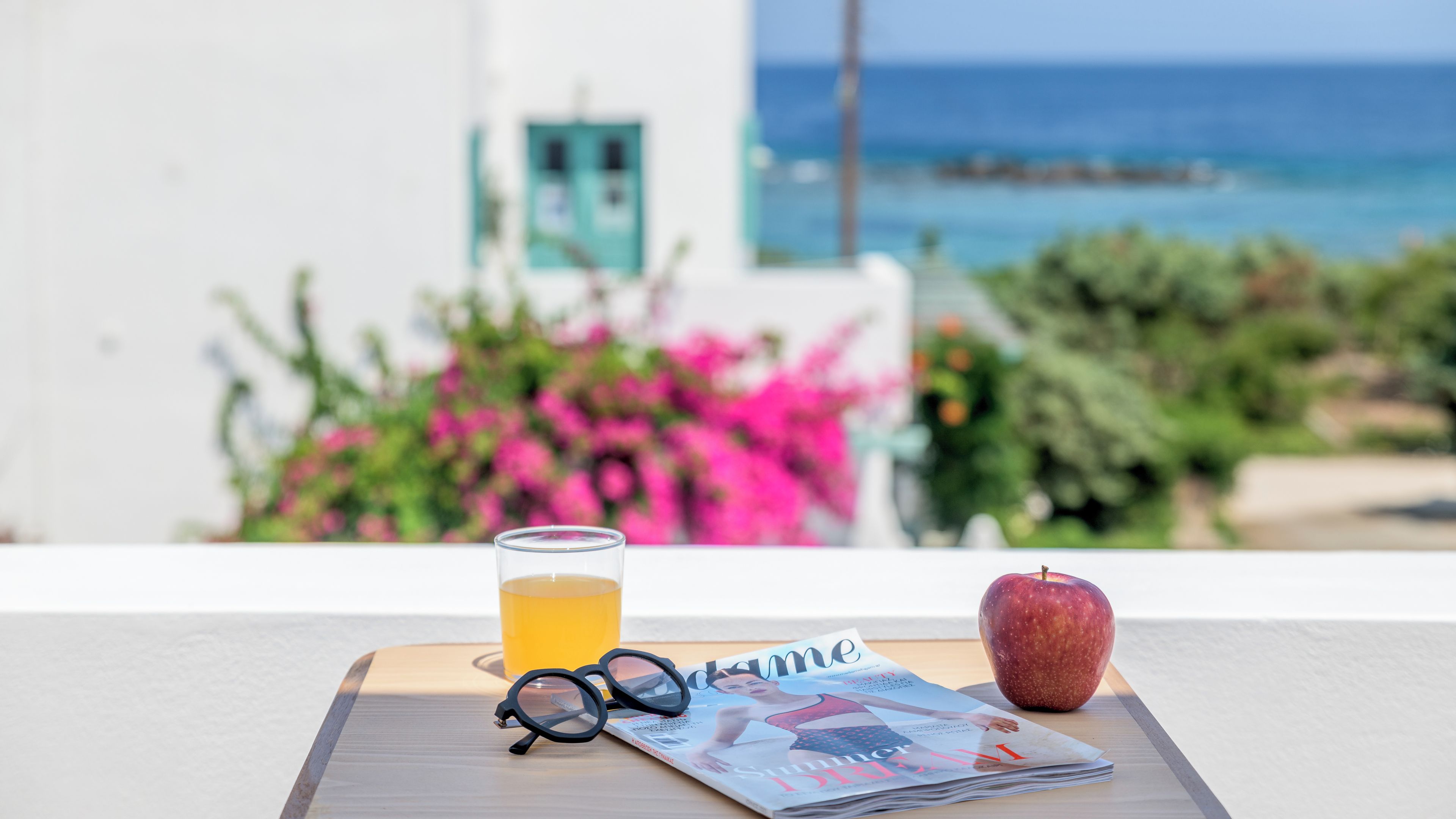 A table with a magazine, a glass of yellow liquid, a pair of glasses, and an apple.
