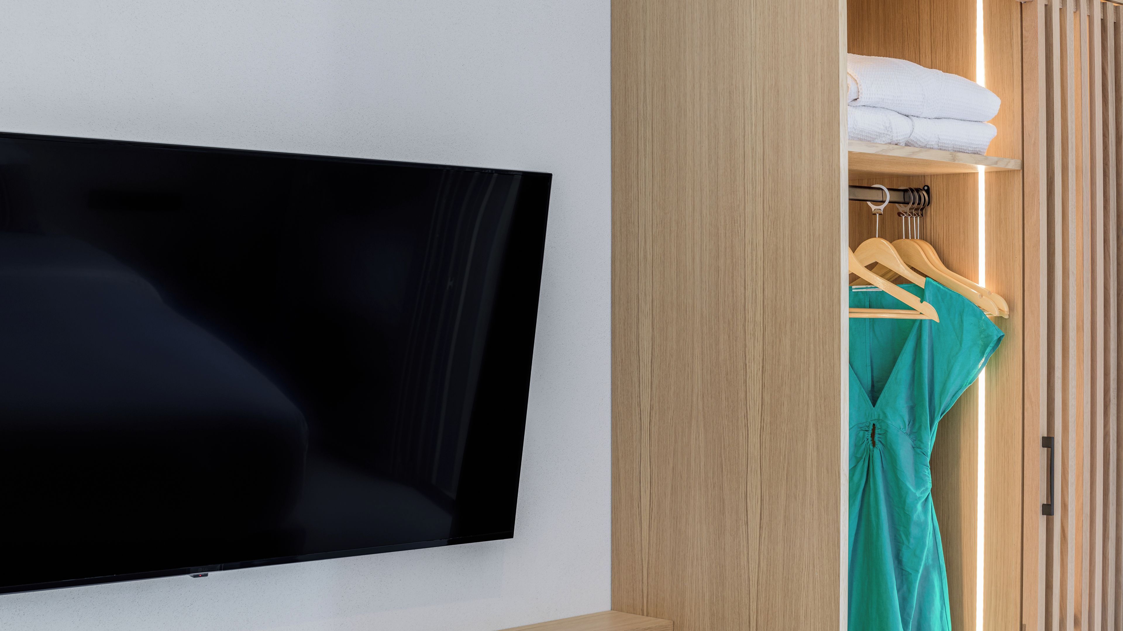 A room with a flat-screen TV mounted on the wall, a wooden wardrobe with a mirror, and a clothing rack with a teal dress and white towels.