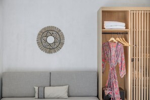 A neutral-toned room with a grey sofa, a wooden wardrobe with a hanging robe, and a circular wall decoration. - Nove Milos Luxury (Milos)