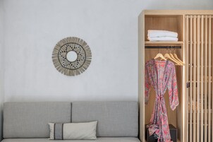 Quarto em tons neutros, com um sofá cinzento, guarda-roupa de madeira com cabides e um ornamento circular na parede.