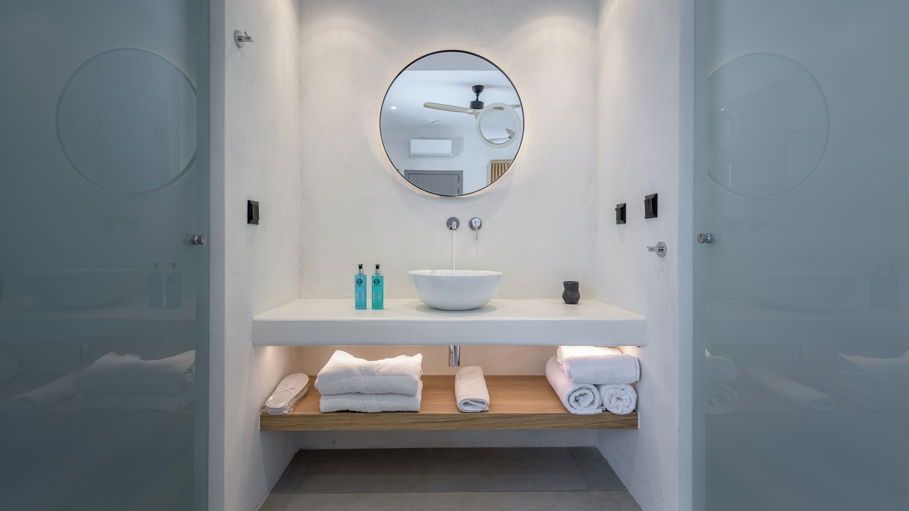 Une salle de bain moderne avec un lavabo blanc, un miroir rond et une étagère en bois où sont posées des serviettes.