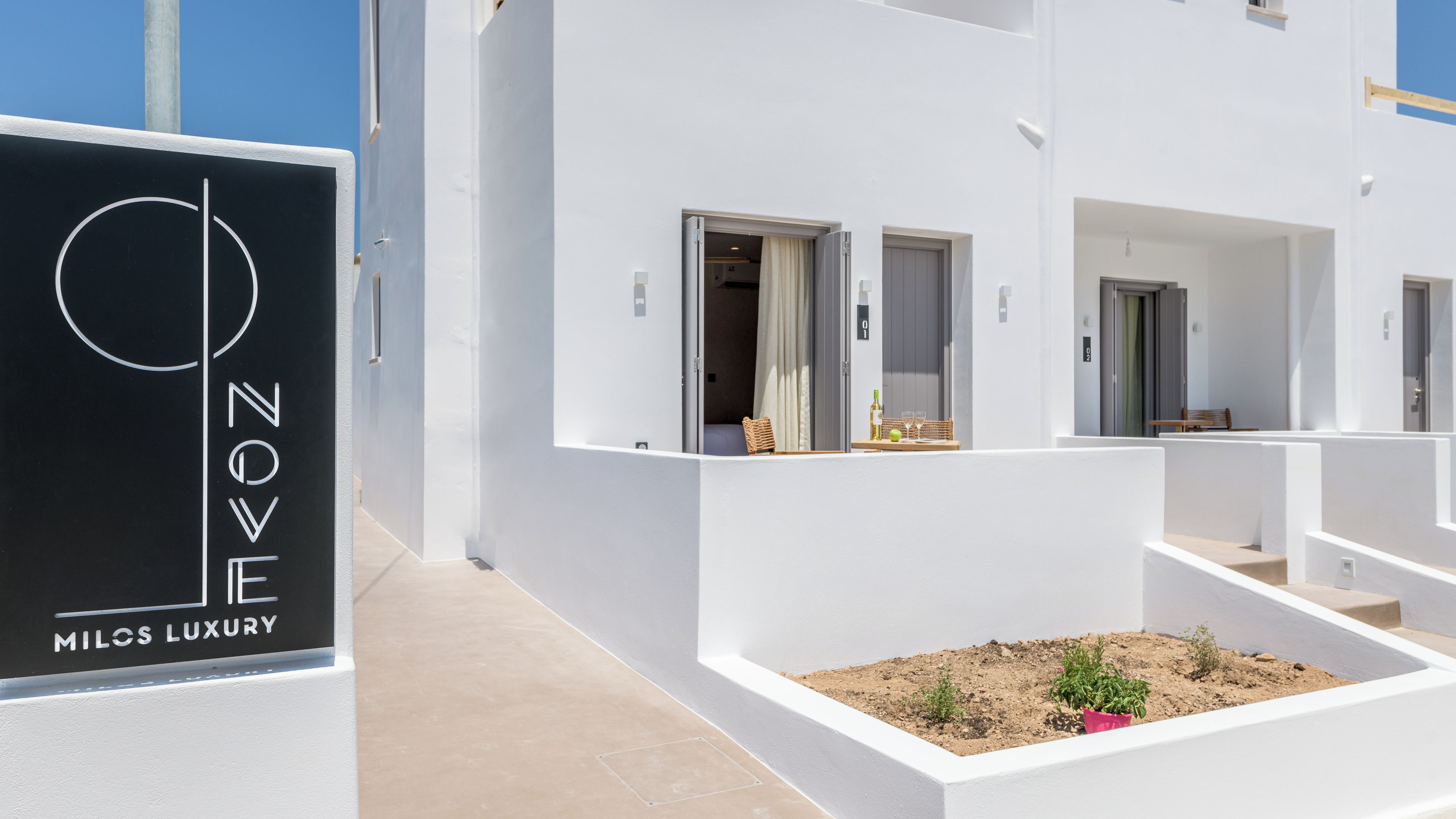 Um edifício moderno com paredes exteriores brancas, uma placa com a inscrição "NOVE MILOS LUXURY" e uma pequena área ajardinada com plantas.
