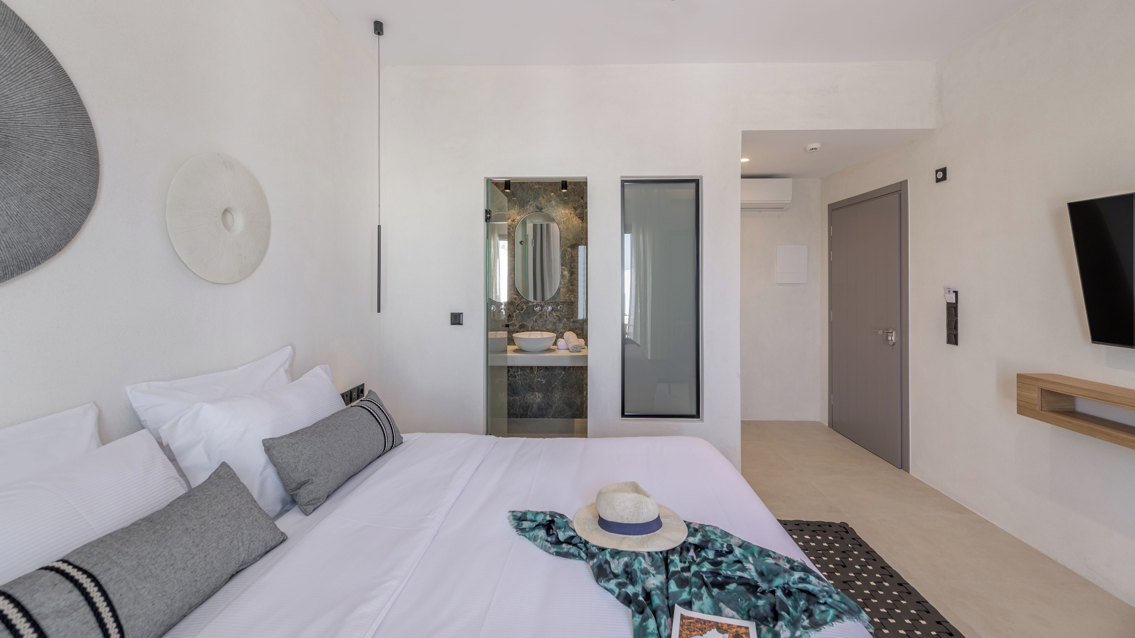 Una camera d'albergo moderna con un letto, uno specchio grande, una TV e un bagno visibile tramite una porta in vetro.
