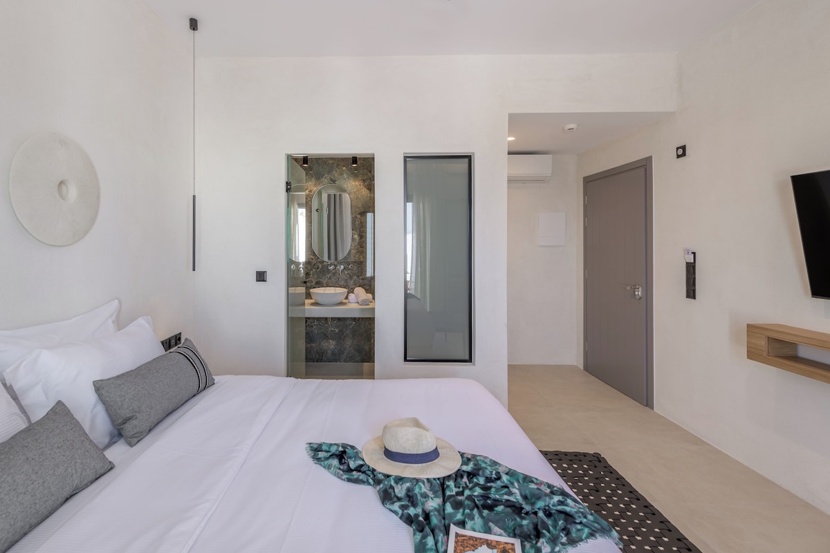 Ein modernes Hotelzimmer mit Bett, einem großen Spiegel, einem Fernseher und einem durch eine Glastür einsehbaren Badezimmer.