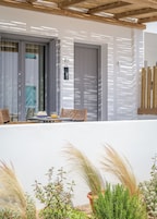Uma área externa iluminada pelo sol com mesas e cadeiras, uma porta com placa de número e plantas em vasos.