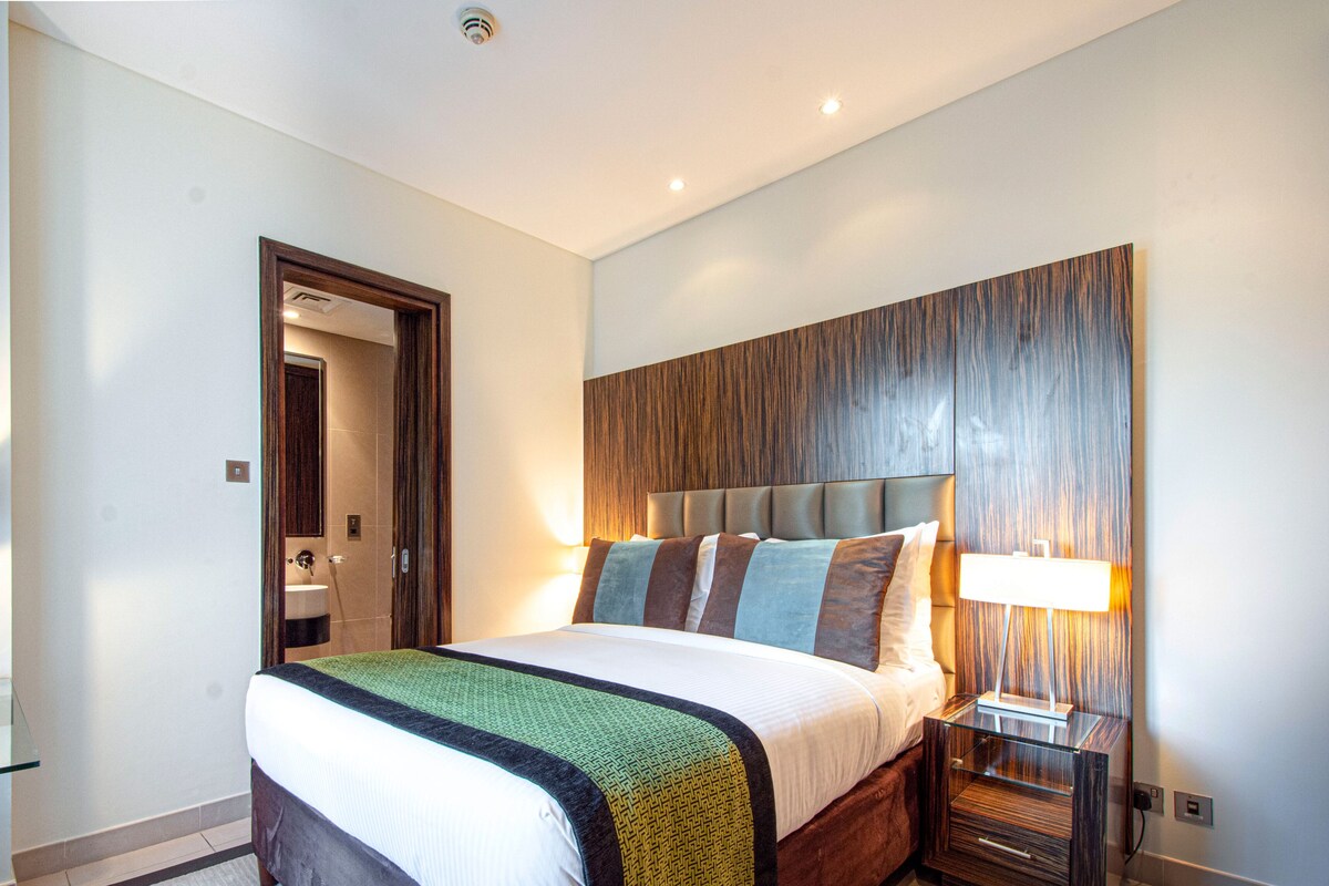 A hotel room with a bed, bedside table, lamp, and a door leading to a bathroom.