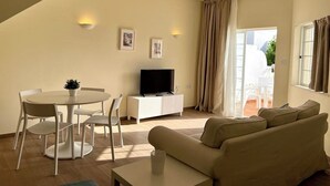 A living room with a sofa, a dining table with chairs, a TV on a white cabinet, and a window with curtains. - Balaia Plaza Al (Albufeira)