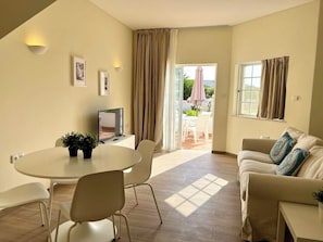 A modern living room with a round table, two chairs, a sofa, and a TV. There is a sliding glass door leading to an outdoor area with a patio table and chairs. - Balaia Plaza Al (Albufeira)