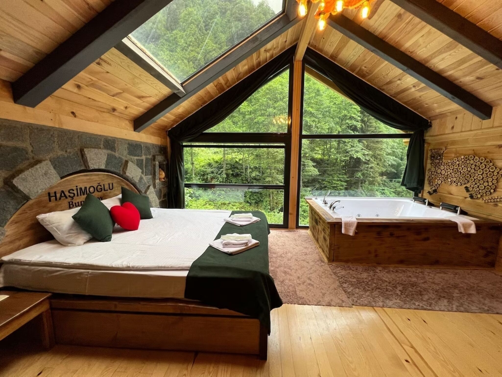 A wooden room with a bed, a bench, and a bathtub. There is a large window with a view of trees.