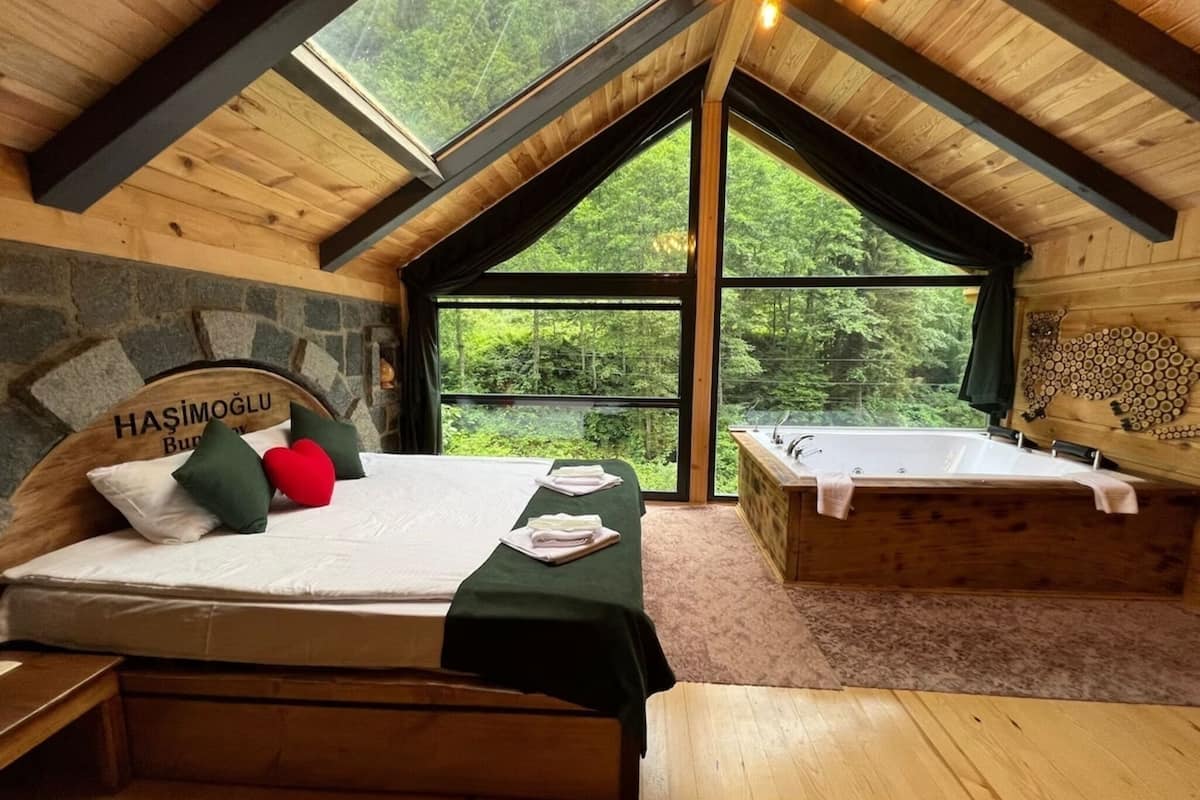 A wooden room with a bed, a bench, and a bathtub. There is a large window with a view of trees.