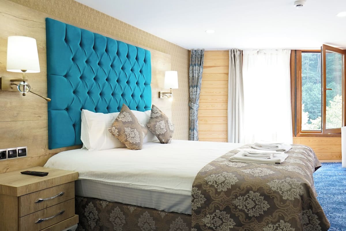 A hotel room with a large bed, a wooden headboard, two bedside lamps, a white armchair, and a window with curtains.