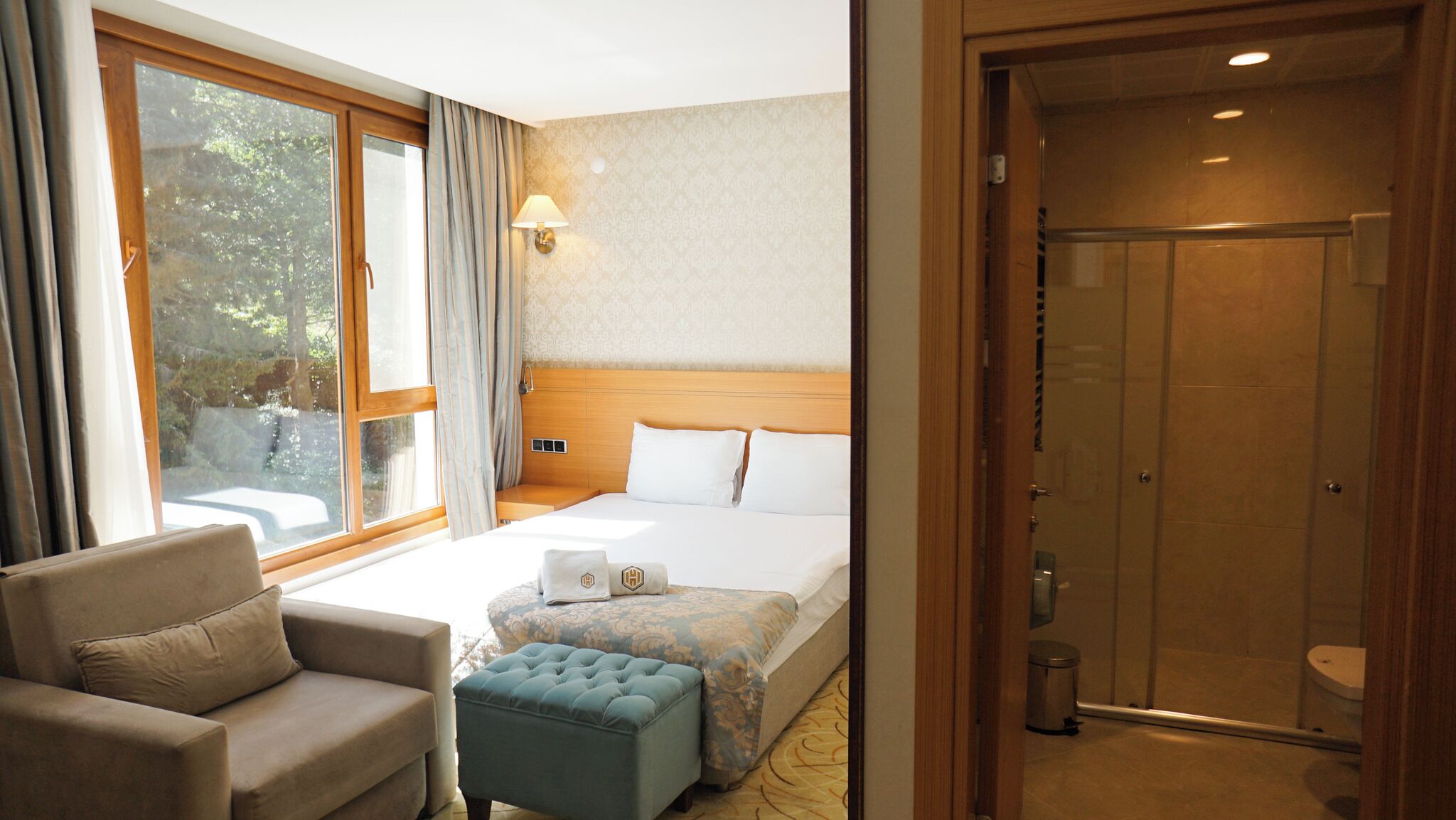 A hotel room with a bed, a sofa, a small ottoman, a nightstand, a lamp, and a shower.