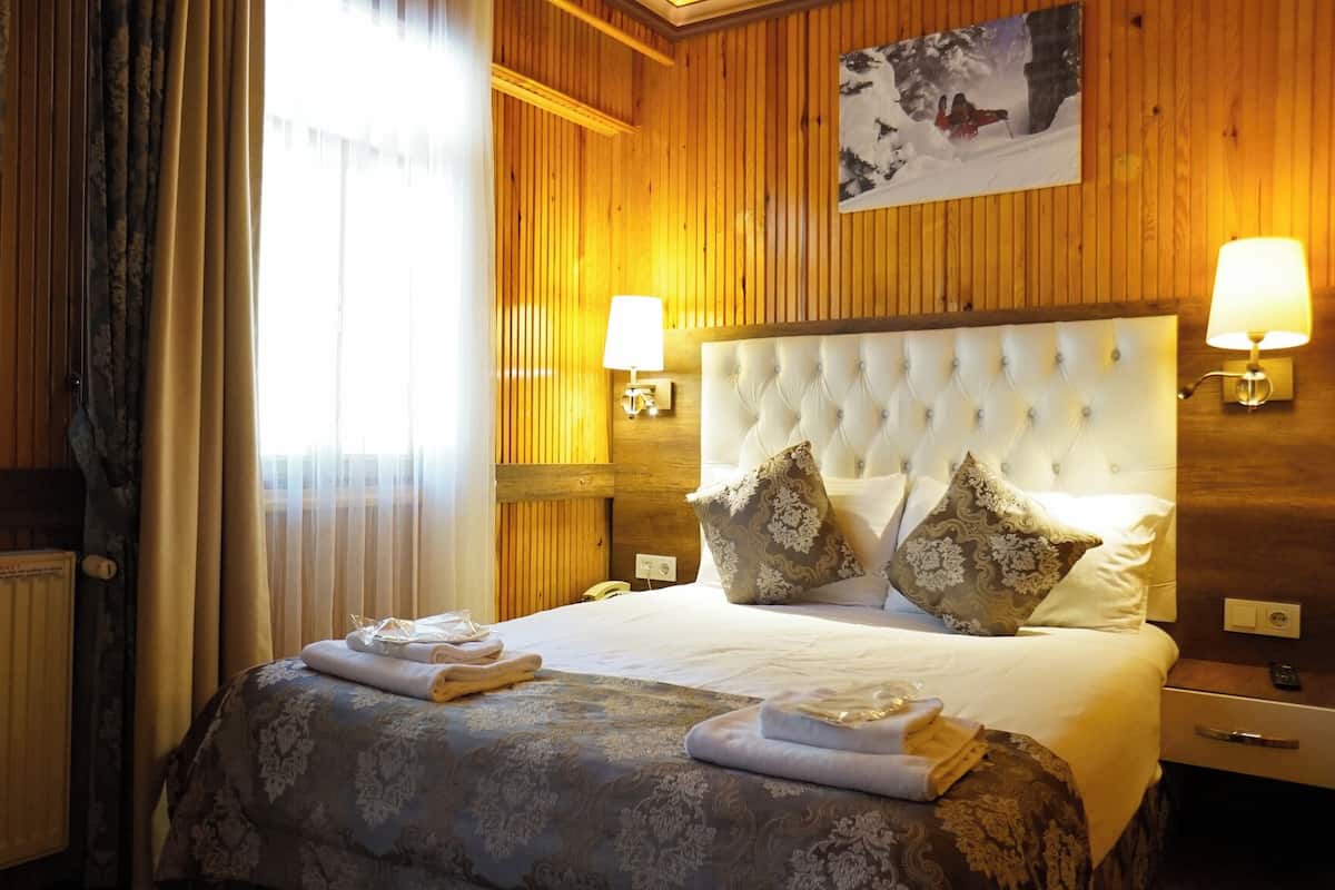 A hotel room with a large bed, two wall-mounted lamps, a painting, and a window with curtains.