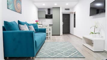 Una sala de estar moderna con un sofá color turquesa, una alfombra estampada, un televisor de pantalla plana y una cocina minimalista al fondo.