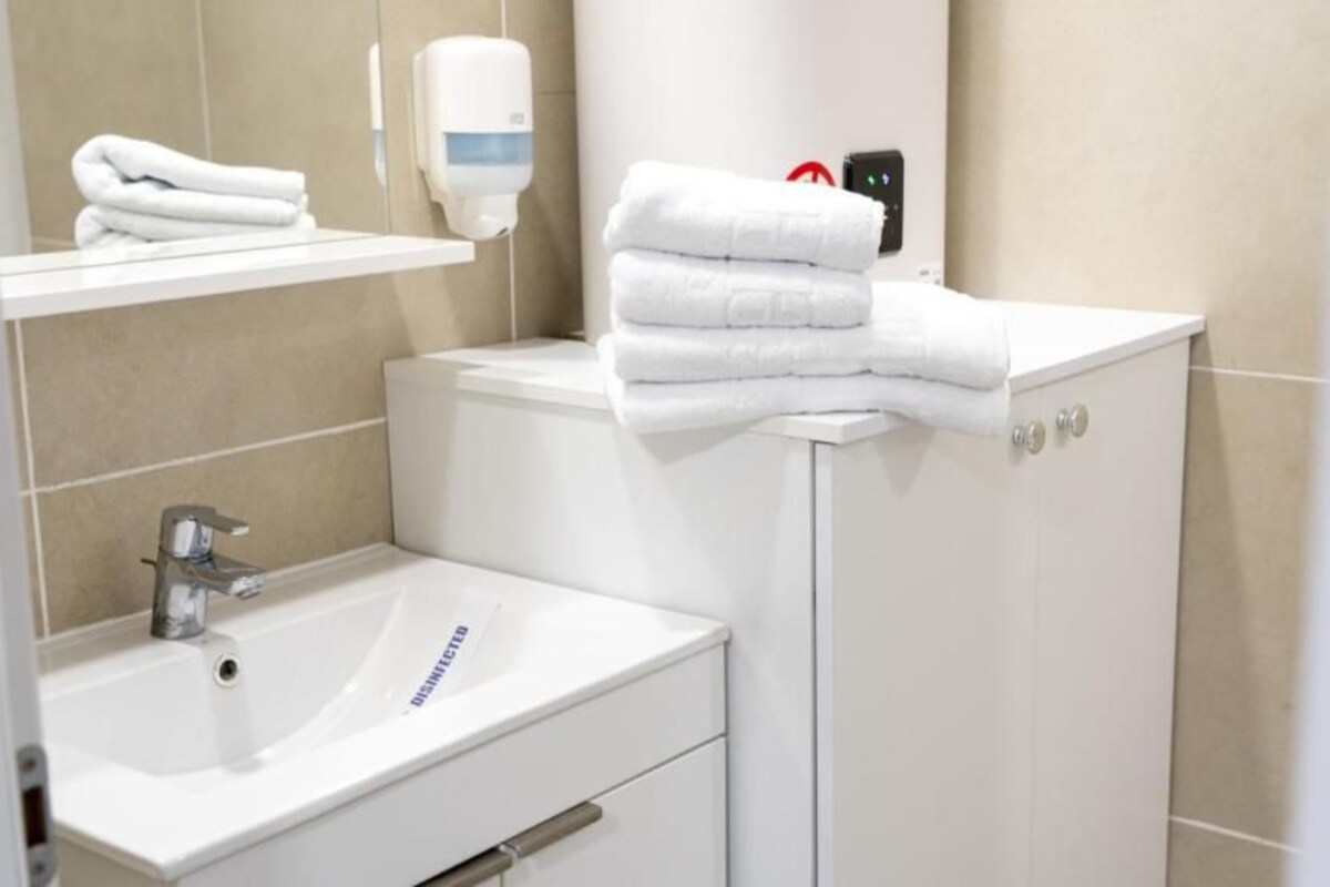 A bathroom with a sink, a mirror, a towel dispenser, and a stack of towels on a cabinet.