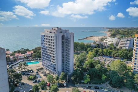 A high-rise hotel with a pool, surrounded by greenery and a beach.
